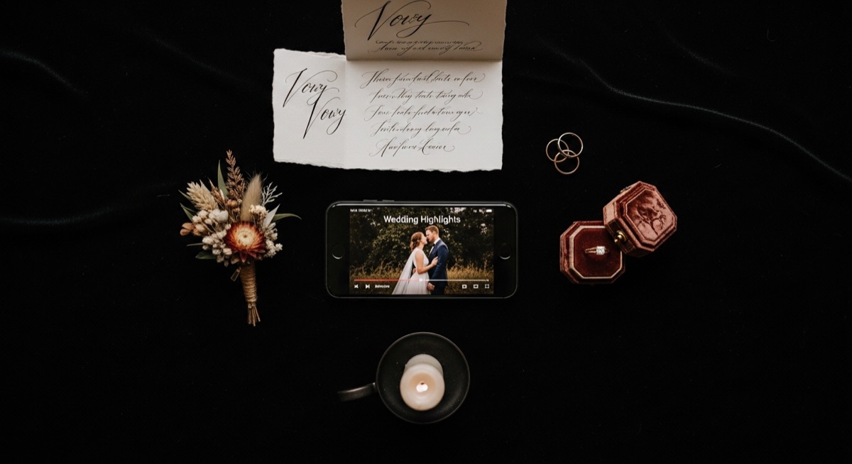 Wedding flat lay with iPhone showing highlights video, boutonniere, rings, vow card, and candle